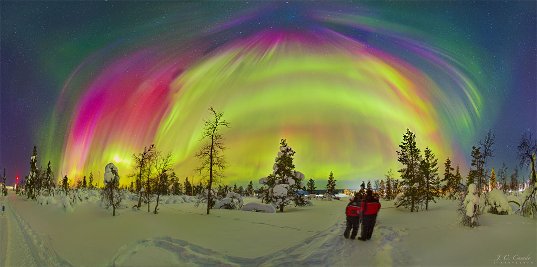 Two people dressed in red coats are standing on a snowy
landscape with bare trees. Above, many aurorae of different 
colors appear, with some stars visible in the background.
Please see the explanation for more detailed information.