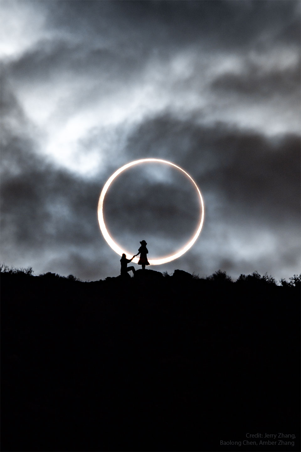 An annular solar eclipse appears in the background with
the dark Moon appearing completely internal to the bright Sun.
In the foreground is a ridge with the silhouettes of two people, 
one standing, and one kneeling. 
Please see the explanation for more detailed information.