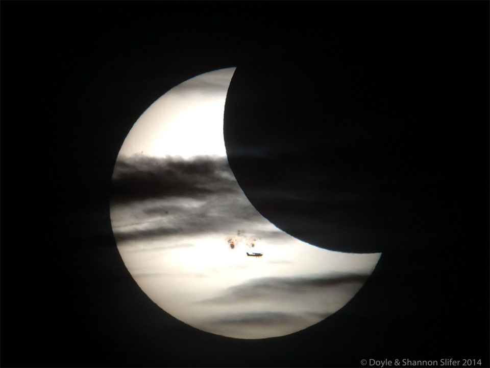 A partially eclipse Sun is shown. In front of the Sun 
are sunspots, the Moon, clouds, and an airplane.
Please see the explanation for more detailed information.