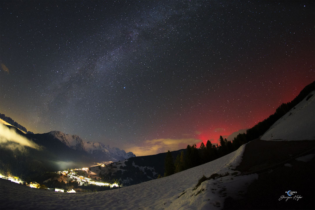 The night sky over a valley is shown complete with 
the central band of the Milky Way Galaxy crossing up from
 the lower left. On the right the sky just over the hill
glows an unusual red: aurora.
Please see the explanation for more detailed information.