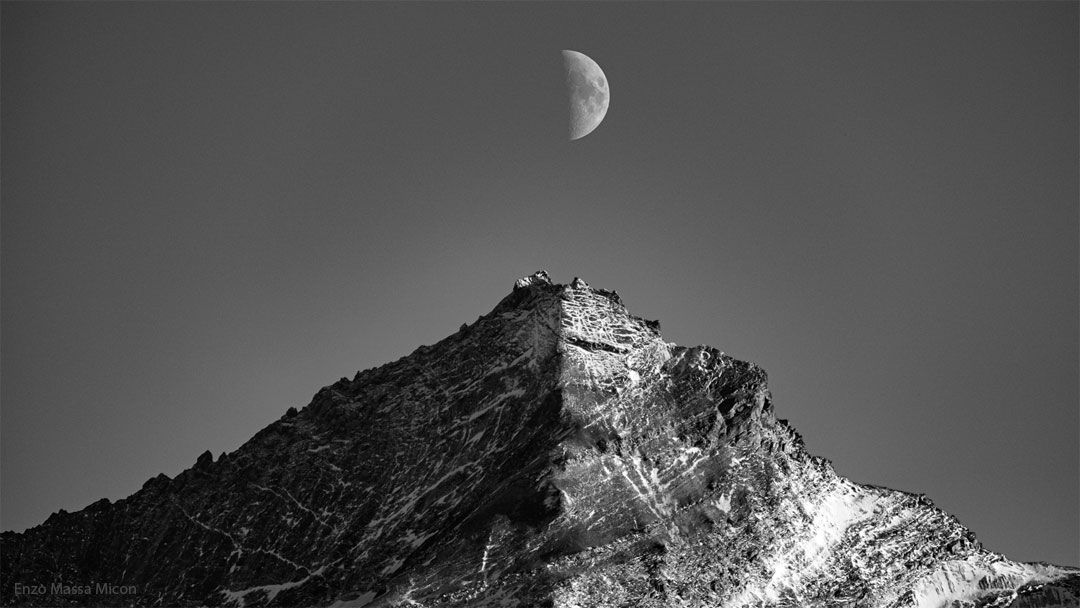 A black and white image of the Moon and a mountain are shown.
Both are half lit by the Sun, with the other half shadowed. The
half-moon is directly above the mountain peak.
Please see the explanation for more detailed information.