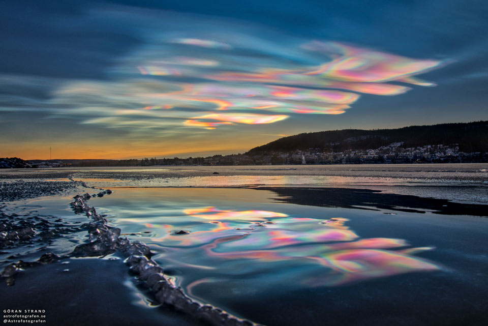 A landscape shows the sky above, shallow water in front,
and hills to the right. The sunset sky is filled with
beautifully multicolored clouds. These clouds are clearly
reflected by the calm water in front. 
Please see the explanation for more detailed information.