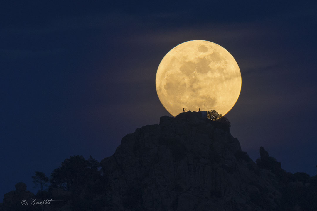 Earth's Moon is shown just beyond a rocky hill. The Moon is
near full phase. On the hill the silhouette of a person looking
through a telescope can be seen. A rollover darkens part of the 
Moon that looks to some like a human face.
Please see the explanation for more detailed information.